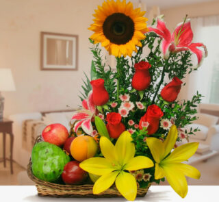 Canasta de mimbre con frutas variadas y arreglo floral con girasol, rosas rojas, lirios amarillos y rosas, sobre una mesa en una sala de estar desenfocada