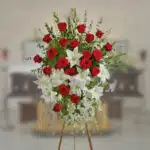 Arreglo floral grande sobre caballete con rosas rojas y lirios blancos en primer plano, con ataúd desenfocado al fondo