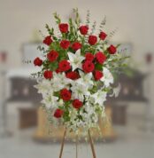 Arreglo floral grande sobre caballete con rosas rojas y lirios blancos en primer plano, con ataúd desenfocado al fondo