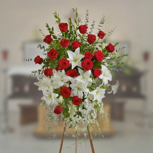 Arreglo floral fúnebre con rosas rojas y lirios blancos Arreglo floral grande sobre caballete con rosas rojas y lirios blancos en primer plano, con ataúd desenfocado al fondo