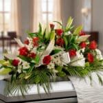 Arreglo floral alargado con rosas rojas, lirios blancos y follaje verde sobre un ataúd en una sala iluminada