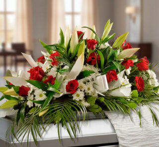 Arreglo floral alargado con rosas rojas, lirios blancos y follaje verde sobre un ataúd en una sala iluminada