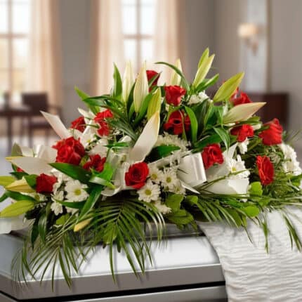 Arreglo floral alargado con rosas rojas, lirios blancos y follaje verde sobre un ataúd en una sala iluminada