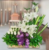 Arreglo floral rectangular con lirios y rosas blancas, lazos morados y follaje verde sobre fondo de sala velatoria