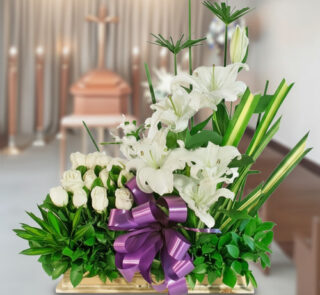 Arreglo floral rectangular con lirios y rosas blancas, lazos morados y follaje verde sobre fondo de sala velatoria