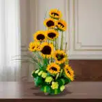 Arreglo floral vertical con girasoles amarillos y flores verdes en base de hojas, colocado sobre una mesa de madera en interior elegante