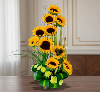 Arreglo floral vertical con girasoles amarillos y flores verdes en base de hojas, colocado sobre una mesa de madera en interior elegante