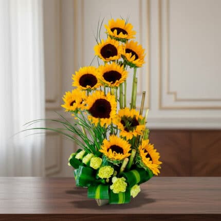 Arreglo floral vertical con girasoles amarillos y flores verdes en base de hojas, colocado sobre una mesa de madera en interior elegante