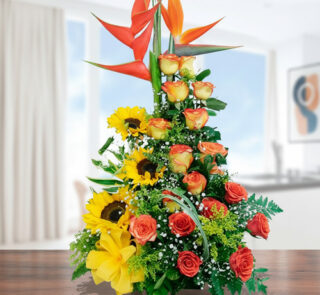 Arreglo floral vertical con rosas naranjas, girasoles, flores amarillas y ave del paraíso sobre una mesa de madera en interior luminoso