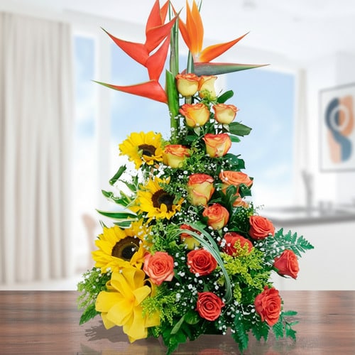 Arreglo floral con rosas, girasoles y ave del paraíso sobre mesa de madera Arreglo floral vertical con rosas naranjas, girasoles, flores amarillas y ave del paraíso sobre una mesa de madera en interior luminoso