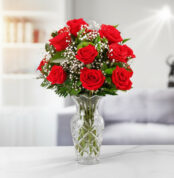 Ramo de rosas rojas con paniculata blanca y hojas verdes en jarrón de cristal tallado sobre mesa blanca en salón moderno desenfocado