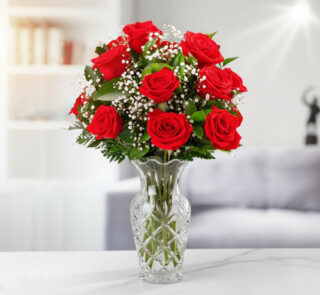 Ramo de rosas rojas con paniculata blanca y hojas verdes en jarrón de cristal tallado sobre mesa blanca en salón moderno desenfocado