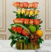 Arreglo de rosas naranjas y amarillas combinado con manzana verde, uvas, naranja y granada sobre base de bambú, colocado en mesa de mármol