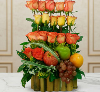 Arreglo de rosas naranjas y amarillas combinado con manzana verde, uvas, naranja y granada sobre base de bambú, colocado en mesa de mármol