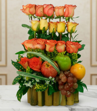 Arreglo de rosas naranjas y amarillas combinado con manzana verde, uvas, naranja y granada sobre base de bambú, colocado en mesa de mármol