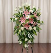 Arreglo floral alto sobre caballete con lirios rosas, rosas blancas y follaje verde frente a una cortina beige