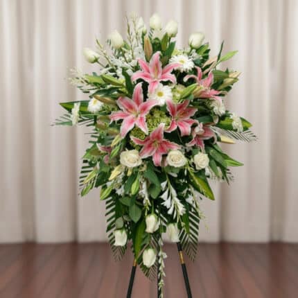 Arreglo floral alto sobre caballete con lirios rosas, rosas blancas y follaje verde frente a una cortina beige