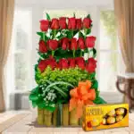 Arreglo floral vertical de rosas rojas con follaje verde en base de bambú y lazo naranja junto a caja de chocolates Ferrero Rocher sobre mesa de madera