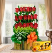 Arreglo floral vertical de rosas rojas con follaje verde en base de bambú y lazo naranja junto a caja de chocolates Ferrero Rocher sobre mesa de madera