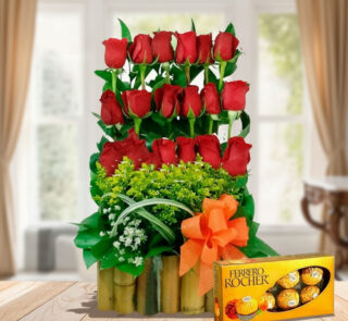 Arreglo floral vertical de rosas rojas con follaje verde en base de bambú y lazo naranja junto a caja de chocolates Ferrero Rocher sobre mesa de madera