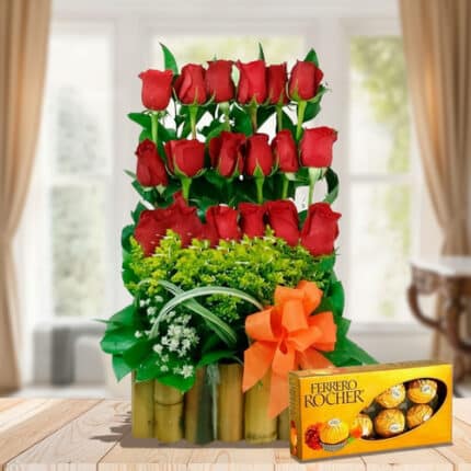Arreglo floral vertical de rosas rojas con follaje verde en base de bambú y lazo naranja junto a caja de chocolates Ferrero Rocher sobre mesa de madera