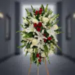 Arreglo floral funerario vertical con lirios blancos, rosas rojas, claveles blancos y follaje verde sobre caballete de madera en pasillo desenfocado