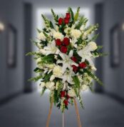 Arreglo floral funerario vertical con lirios blancos, rosas rojas, claveles blancos y follaje verde sobre caballete de madera en pasillo desenfocado