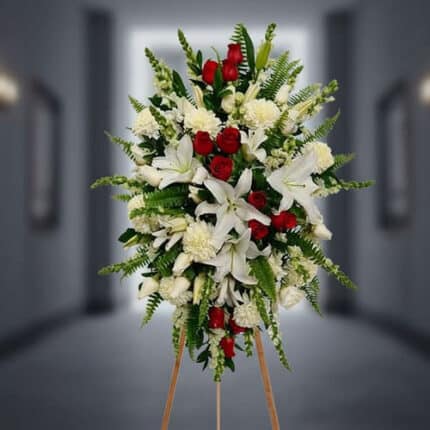 Arreglo floral funerario vertical con lirios blancos, rosas rojas, claveles blancos y follaje verde sobre caballete de madera en pasillo desenfocado