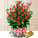 Arreglo floral de rosas rojas con lirios rosas al frente, globo con mensaje de amor y caja de chocolates Ferrero Rocher sobre fondo claro