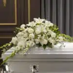 Arreglo de flores blancas con lirios y claveles sobre un ataúd blanco en una sala funeraria elegante