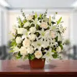 Arreglo floral grande con lirios y rosas blancas en maceta marrón sobre mesa de madera, con fondo desenfocado de oficina