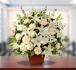 Arreglo floral grande con lirios y rosas blancas en maceta marrón sobre mesa de madera, con fondo desenfocado de oficina