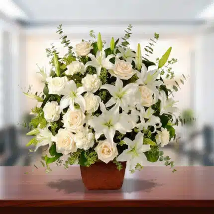 Arreglo floral grande con lirios y rosas blancas en maceta marrón sobre mesa de madera, con fondo desenfocado de oficina