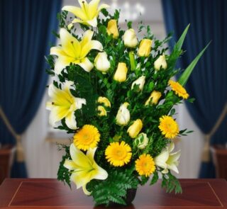 Arreglo vertical de flores amarillas y blancas con lirios, rosas y gerberas sobre una mesa de madera, con cortinas azules desenfocadas al fondo