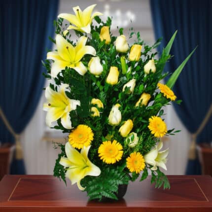 Arreglo vertical de flores amarillas y blancas con lirios, rosas y gerberas sobre una mesa de madera, con cortinas azules desenfocadas al fondo