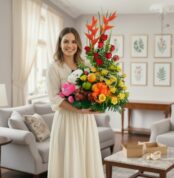 Mujer joven de vestido claro sosteniendo un gran arreglo de flores y frutas con peluche en una sala de estar luminosa