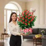 Mujer joven sonriendo sostiene un gran arreglo de rosas rojas y lirios rosados con un globo en forma de corazón que dice Te Amo en una sala elegante
