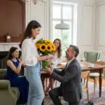 Hombre de traje arrodillado entregando un ramo de girasoles a una mujer joven en un salón luminoso, con dos mujeres sentadas al fondo observando y sonriendo