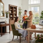Hombre entregando un ramo de flores coloridas a una mujer de pie junto a otra mujer sentada en una oficina elegante y luminosa