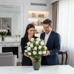 Hombre y mujer sonrientes acomodando un gran ramo de rosas blancas en un jarrón de cristal sobre una mesa en un salón moderno y luminoso