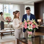 Hombre y mujer observan un gran ramo de flores rosas y blancas sobre una mesa de cristal en una sala de estar moderna y luminosa, con otra pareja al fondo sosteniendo un arreglo floral similar