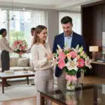 Hombre y mujer observan un gran ramo de flores rosas y blancas sobre una mesa de cristal en una sala de estar moderna y luminosa, con otra pareja al fondo sosteniendo un arreglo floral similar