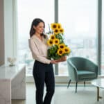 Mujer joven de pie en sala luminosa con ventanal grande, sosteniendo un arreglo floral alto de girasoles amarillos en maceta rectangular de madera