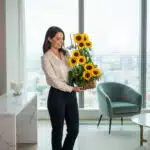 Mujer joven de pie en sala luminosa con ventanal grande, sosteniendo un arreglo floral alto de girasoles amarillos en maceta rectangular de madera