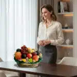 Mujer joven de pie junto a una mesa de madera con una canasta de frutas surtidas, incluyendo manzanas, uvas, peras y cítricos, en una oficina moderna con estantería al fondo