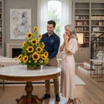 Hombre entregando un gran arreglo de girasoles a una mujer emocionada en una sala de estar elegante con mesa redonda de mármol