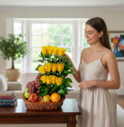 Mujer joven junto a una canasta de mimbre con rosas amarillas en varios niveles y frutas frescas sobre una mesa de madera en sala iluminada