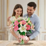 Hombre y mujer sonrientes abrazados mientras sostienen un gran ramo de gerberas rosas, rosas melocotón y lirios blancos en un jarrón de cristal sobre una mesa de madera clara