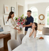 Hombre entregando a una mujer un ramo de lirios rosados mientras otra mujer sentada en la mesa de mármol observa sonriente en un comedor luminoso