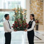 Dos empleados de hotel con uniforme formal sostienen un gran arreglo floral con rosas rojas y aves del paraíso en el lobby luminoso de un edificio